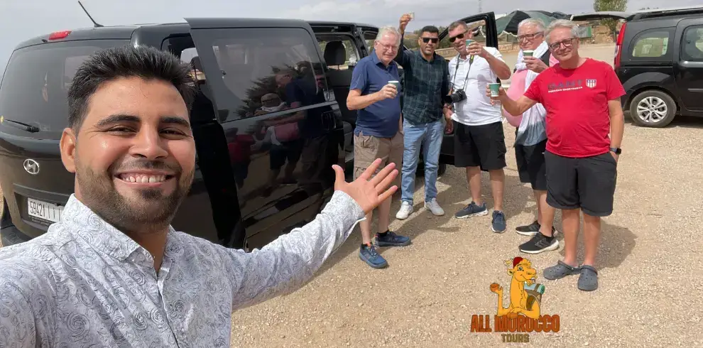 smiling guide taking a selfie with happy travelers holding up drinks next to their van, on a 6 Days Start from Marrakech to Desert to the Moroccan desert.