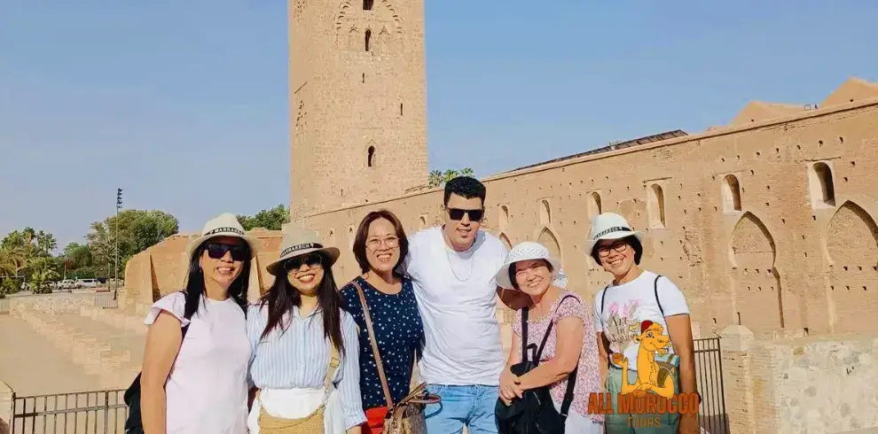 A group of happy tourists posing in front of the tall Koutoubia Mosque tower in Marrakech during a sunny day on their 3 days trip from Marrakech to Fes.