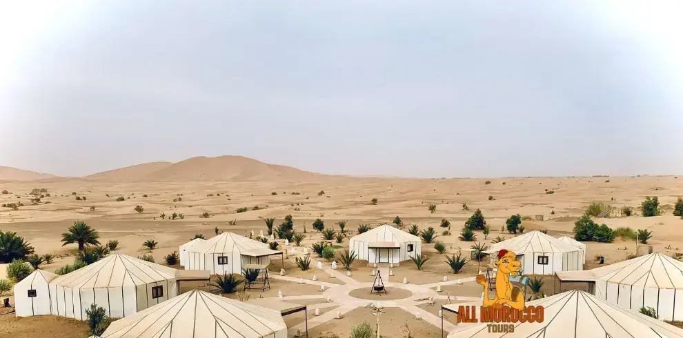 Aerial view of a desert camp in Merzouga with white traditional tents arranged in a symmetric pattern surrounded by sparse vegetation and rolling sand dunes a serene escape on the 3-day journey from Fes to Merzouga.