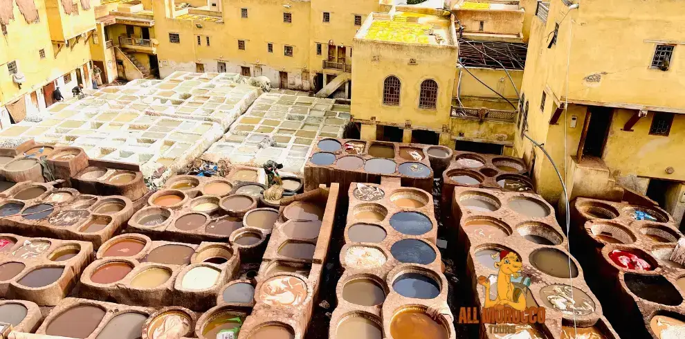 An overhead view of the bustling Fez tannery with workers dyeing leather in vibrant vats, surrounded by the ochre-hued buildings of the medina, a cultural highlight of the 2-day excursion from Fes to the desert.