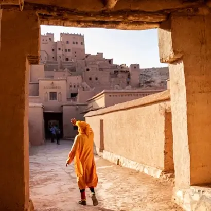 local in traditional attire walking through the ancient alleys of Kasbah Ait Ben Haddou, with the iconic earthen clay architecture of the Errachidia Archives visible in the background under a clear sky