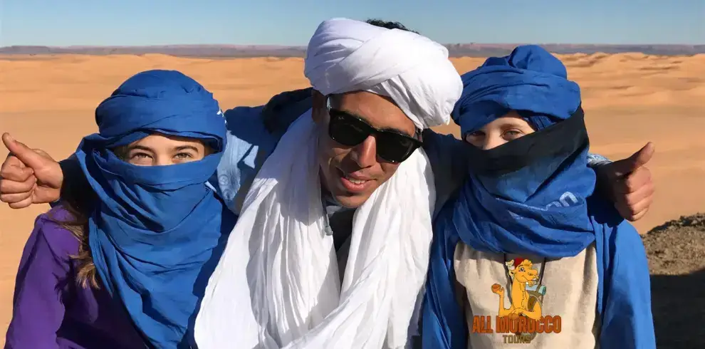 Three happy people with blue scarves around their heads stand in a big sandy desert. They are on a 10-day trip from Casablanca.