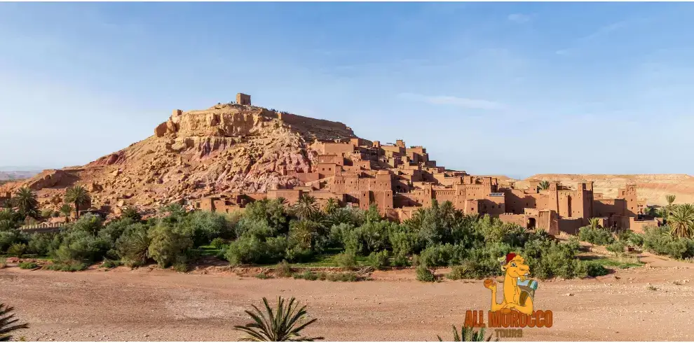 View of the ancient village Ait Ben Haddou with its brown mud buildings and green palm trees, under a clear blue sky. This is a popular stop on a 5 days tour from Casablanca.