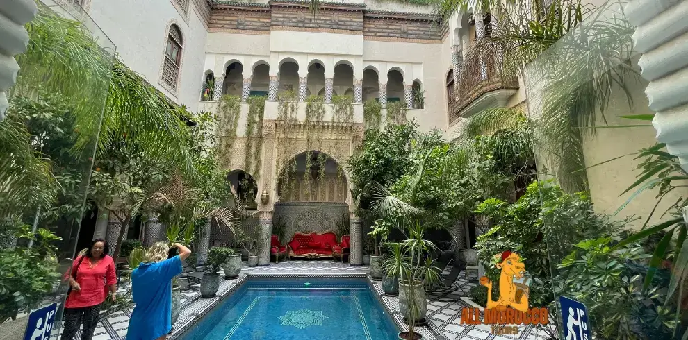 Visitors enjoying the tranquil ambiance of a traditional Riad courtyard in Fes featuring a central poo ornate arches, and lush greenery a restful overnight stay on the 3 Days tour from Fes to sahara desert.