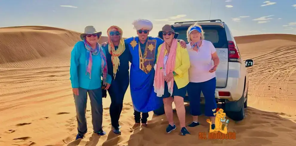 A group of cheerful tourists with a guide in traditional attire next to an off-road vehicle, enjoying the sand dunes on their 7 Days Tour From Marrakech.