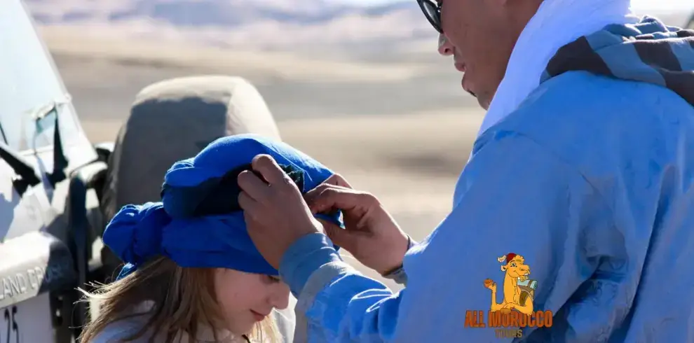 our local guide in traditional blue attire wraps a blue headscarf around a young tourist against the backdrop of the desert, symbolizing cultural experiences on a 12 days tour from Tangier to Marrakech