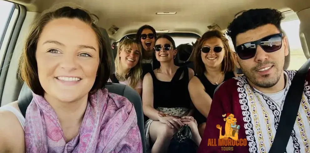 Our guide wearing traditional Moroccan attire poses for a selfie with five smiling women from the USA, all seated comfortably in a vehicle during a 4 days tour from Casablanca Morocco.