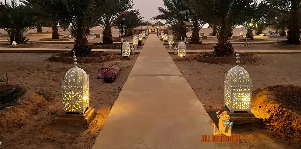 path lined with lit lanterns and palm trees at dusk leading to tents in a desert camp ready for an overnight stay on a 2 Days Tour From Marrakech To Fes.